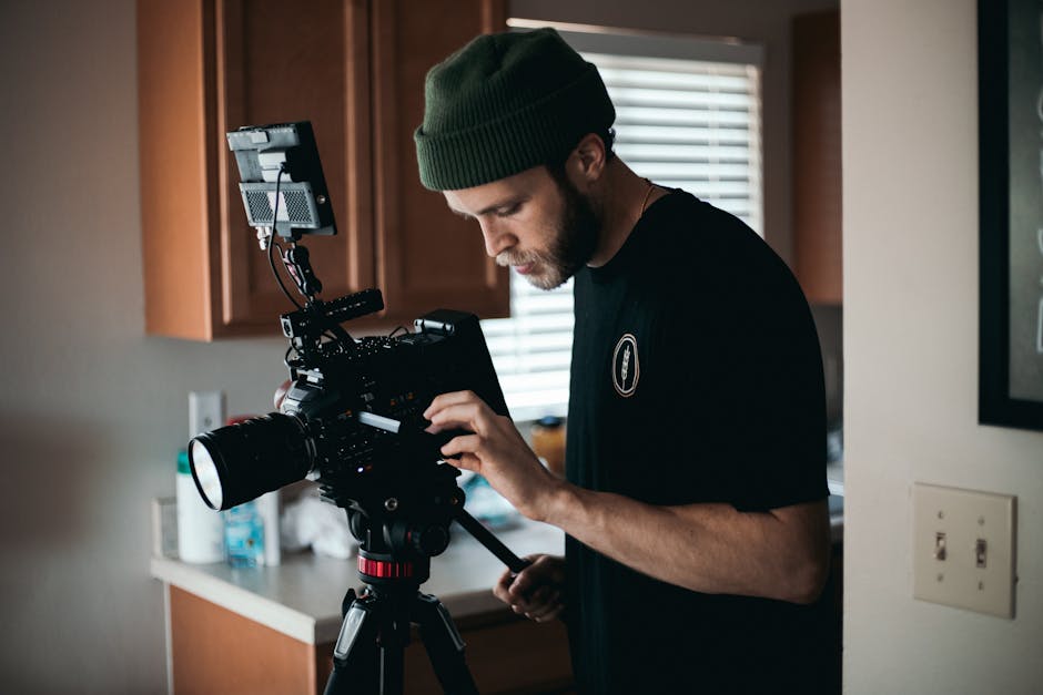 A bearded man with a beanie operates camera equipment indoors, focusing intently.